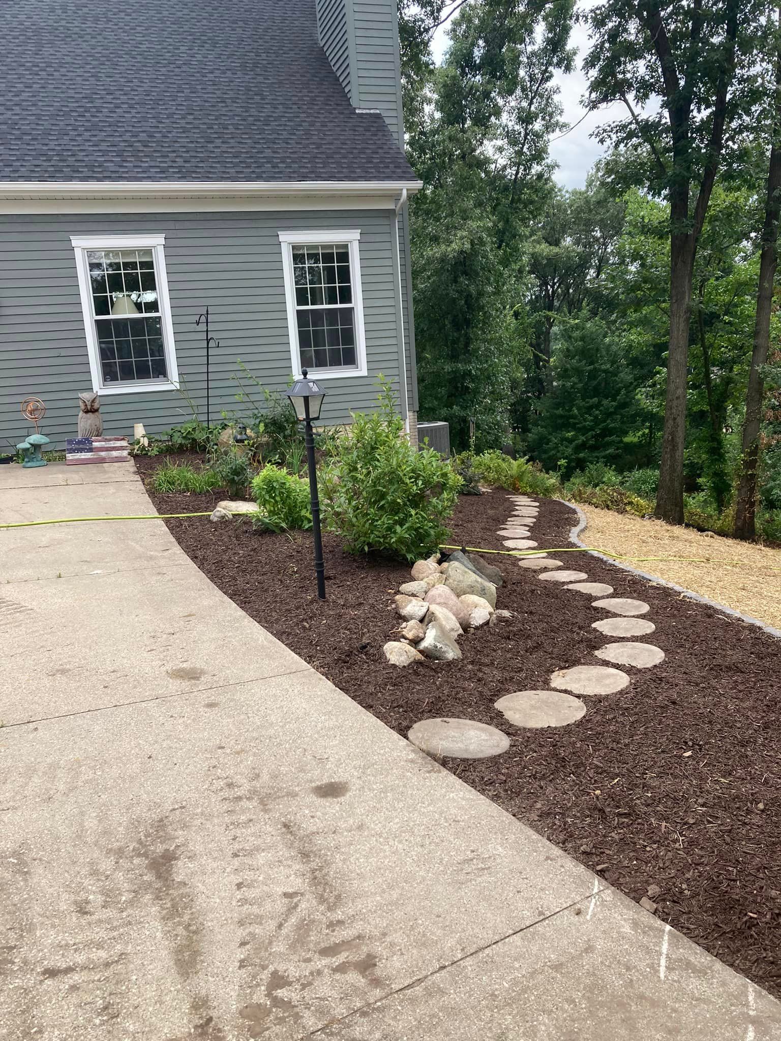 Concrete driveway leading to a house with a garden bed, stepping stones, and trees.