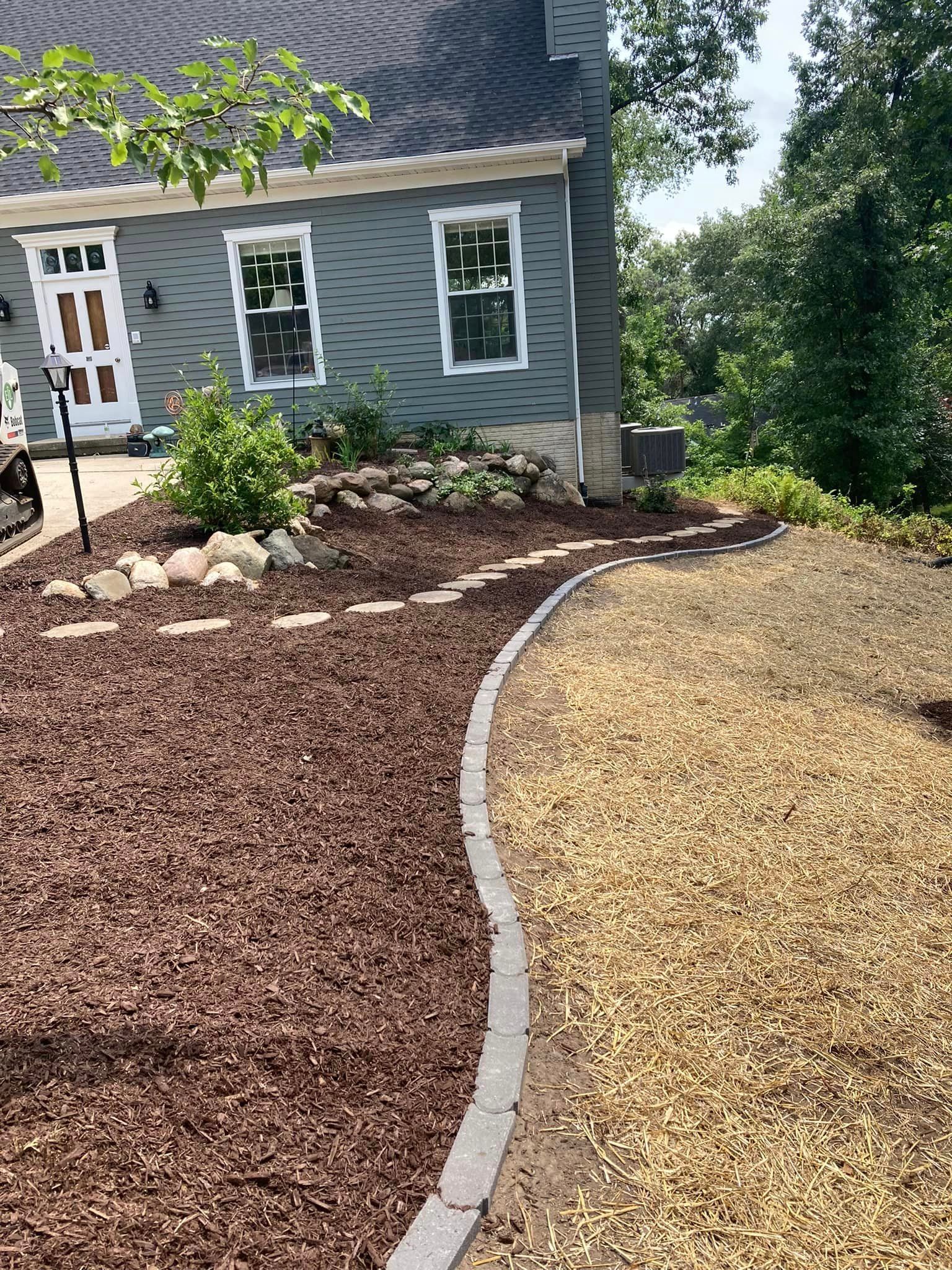 Mulched garden bed next to gravel path, bordered by stones, in front of a blue house.