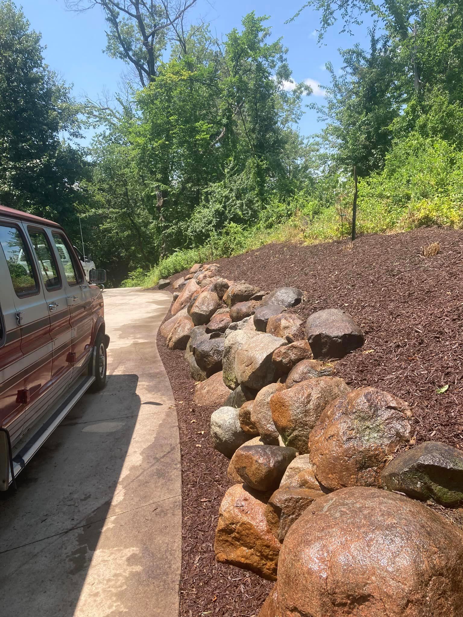 Van parked on concrete next to rock retaining wall on a hill covered in mulch, trees in background.