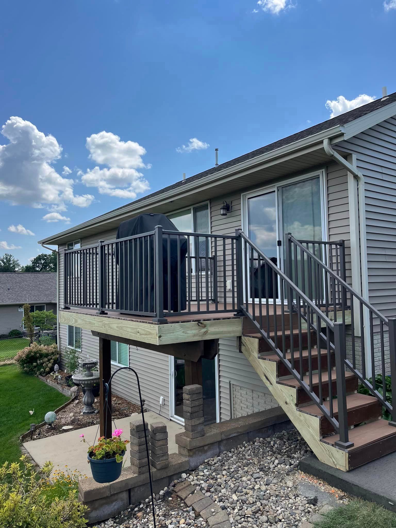 Deck with black railing, stairs, and grill against a two-story house with siding, under a blue sky.