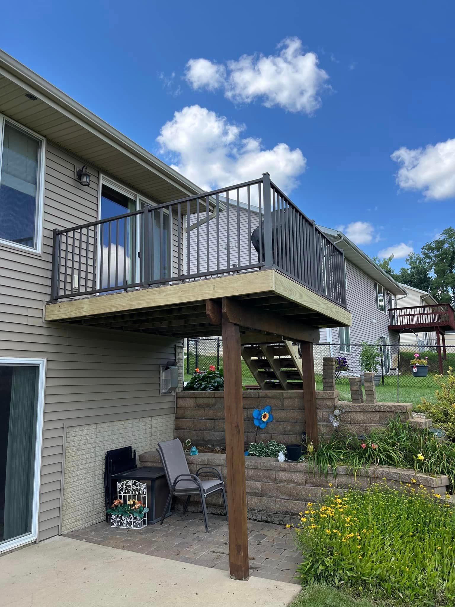 A two-story house with a wooden deck and black railing. A staircase leads to the yard.
