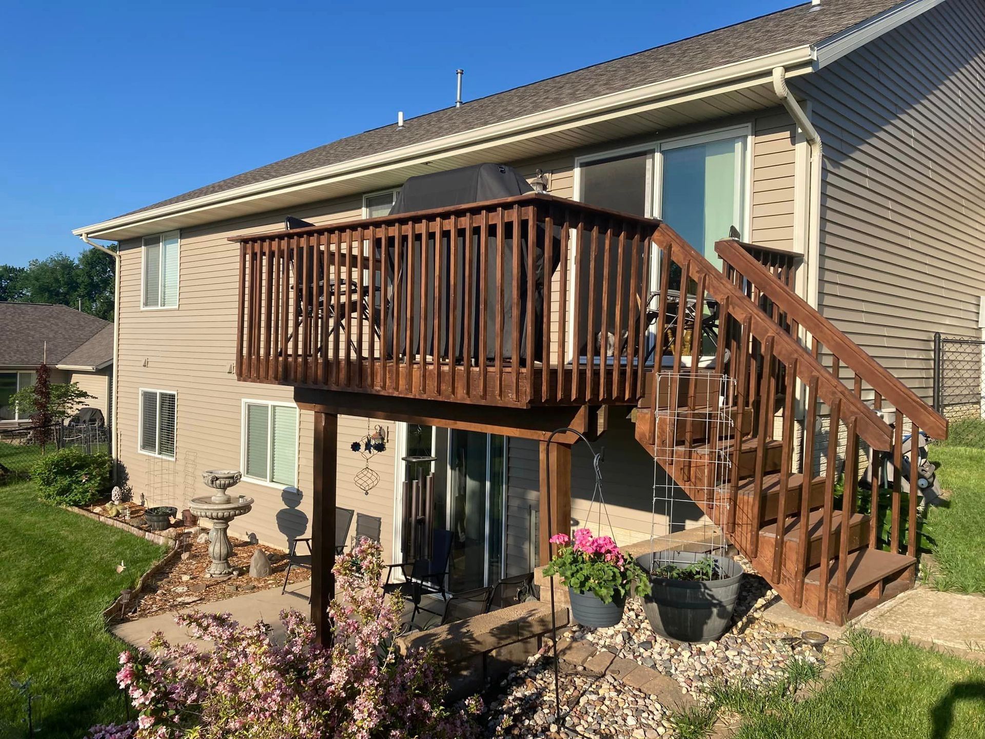 Two-story house with wooden deck and stairs. Beige siding, brown deck stain, green lawn.