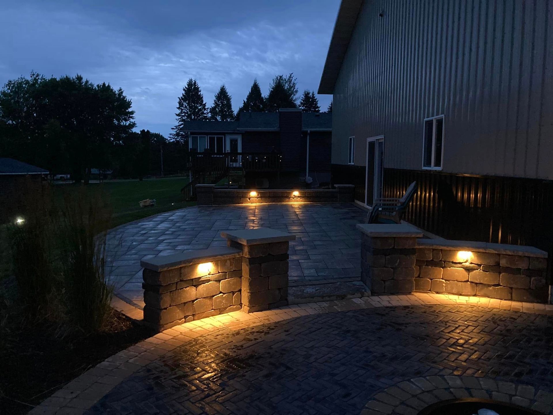 Lit patio at dusk with brick pillars and built-in lights, curving toward a house with a dark sky.