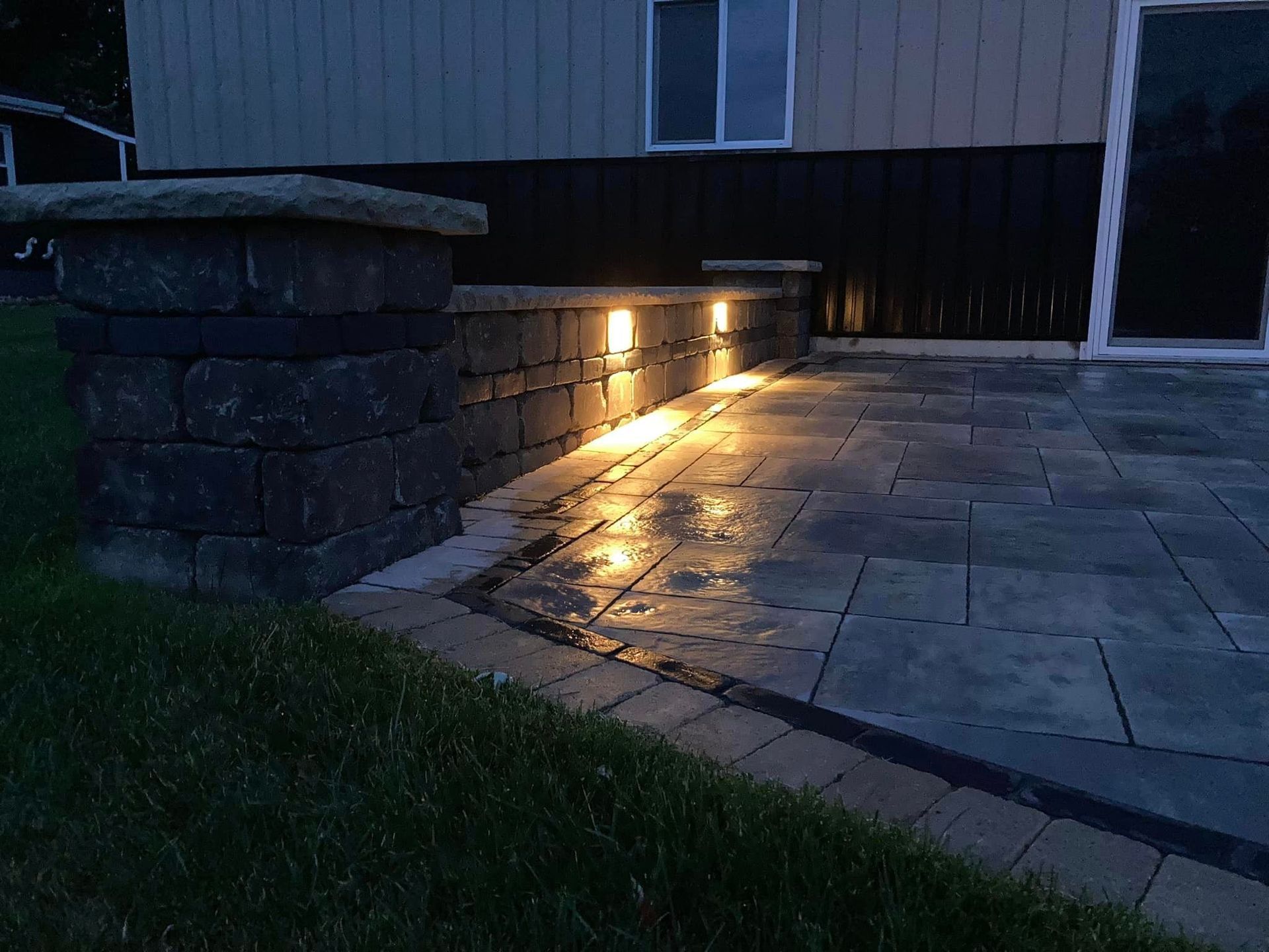 Lit stone patio with built-in wall lights. Dark stone building in the background. Grass in foreground.