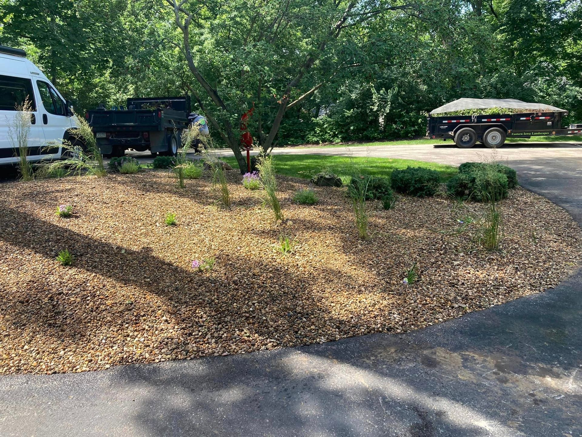Landscaped bed with tan gravel, new plantings, and a central tree in a driveway. Trailers and van visible.
