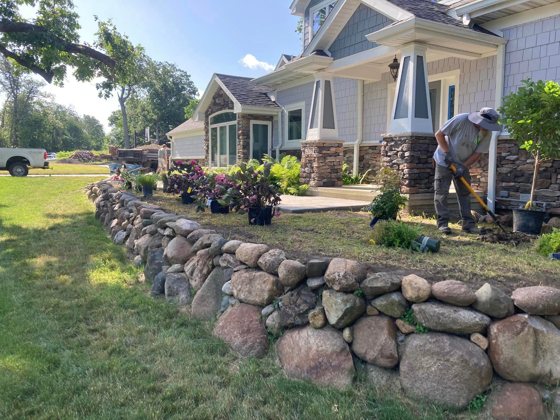 Man gardening near a stone wall and house with blue and gray accents, sunny day.