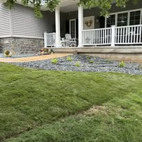 Lush green lawn in front of a house with a white porch and gray stone foundation. Gravel landscaping visible.