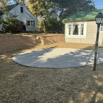 Circular concrete patio in yard with two-story house and retaining wall in the background.