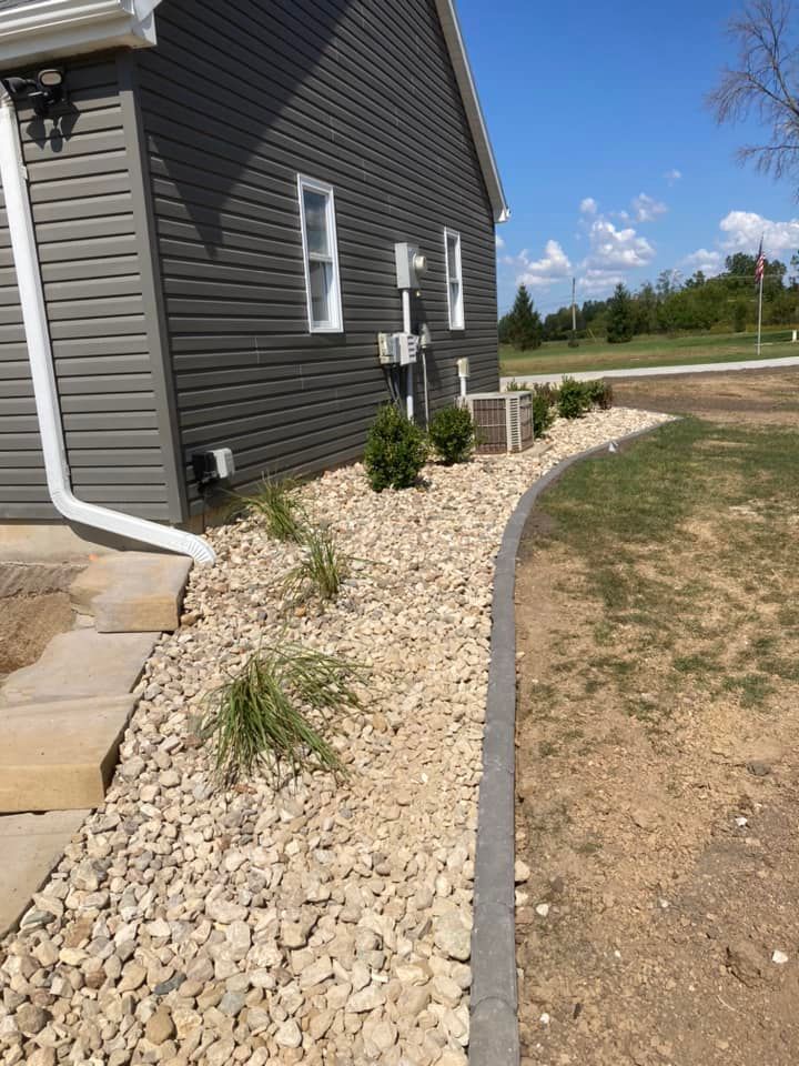 Gray house with rock landscaping, bordering a dirt pathway and green grass.