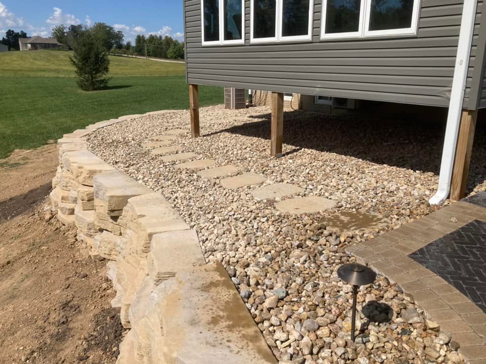 Stone retaining wall and gravel path under a gray house with windows, grass lawn in the background.