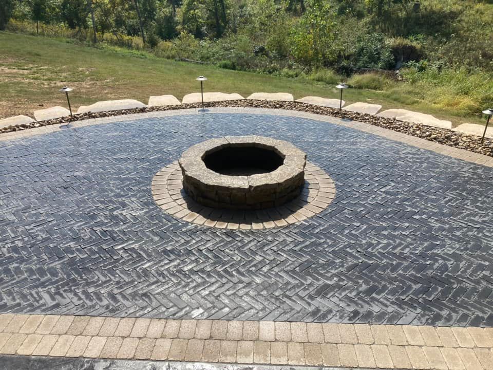 Brick patio with a fire pit, surrounded by landscaping and lights, overlooking a grassy area.