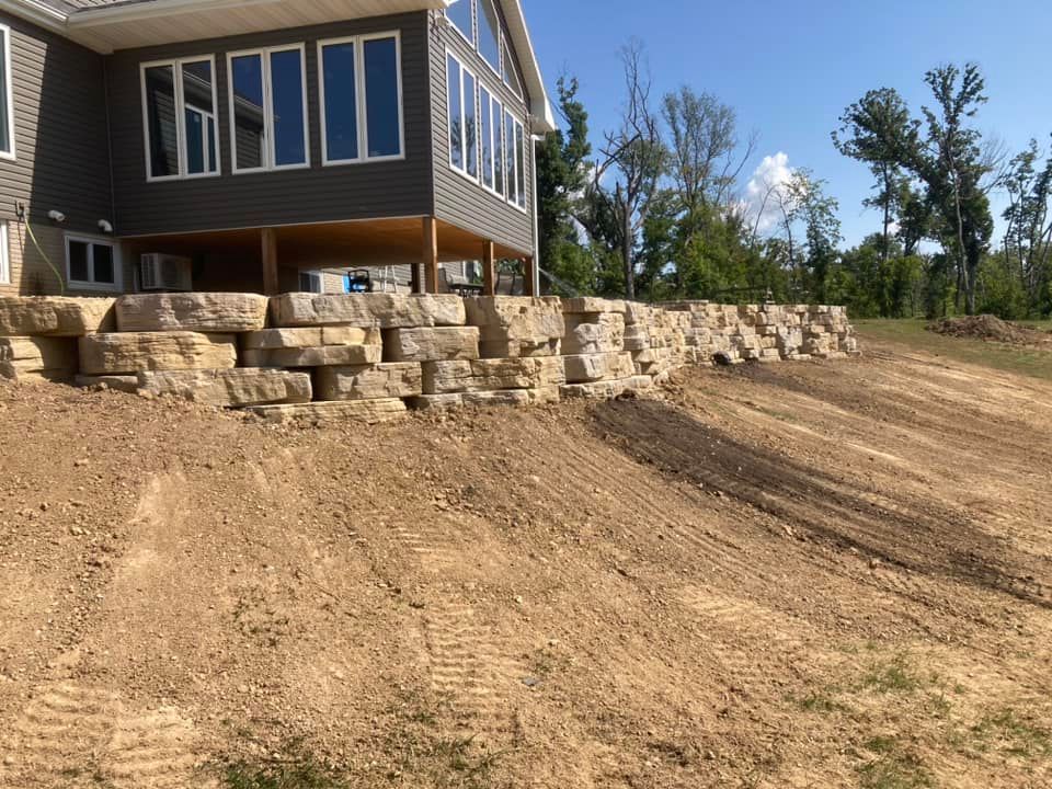 Stone retaining wall supports a house with large windows, on a graded dirt lot, sunny day.