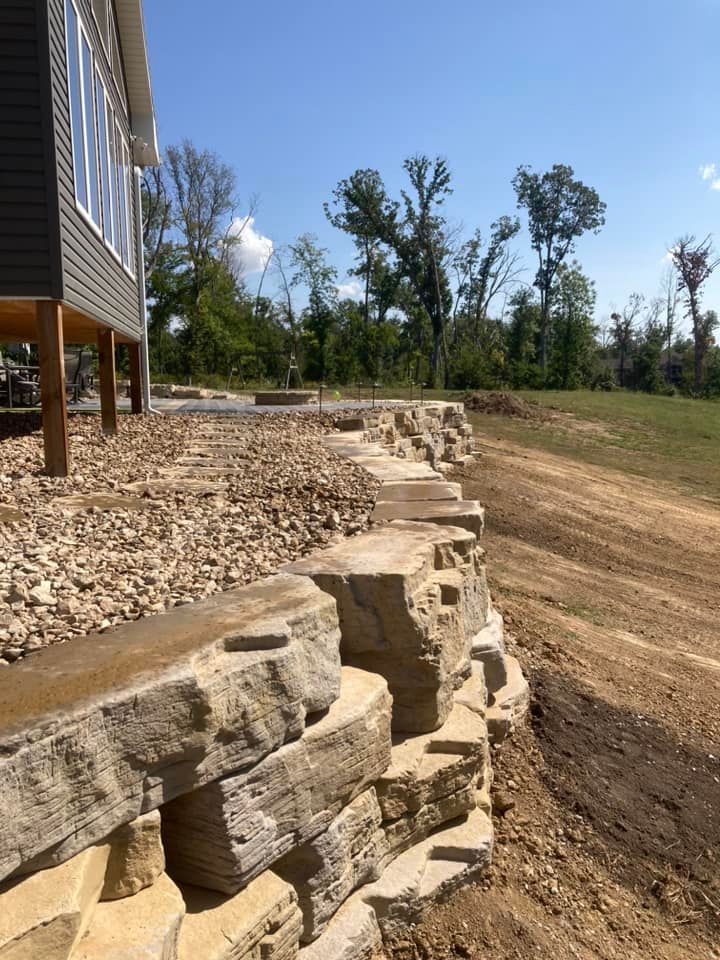 Stone retaining wall along a house with gravel and dirt ground. Blue sky with trees in the background.