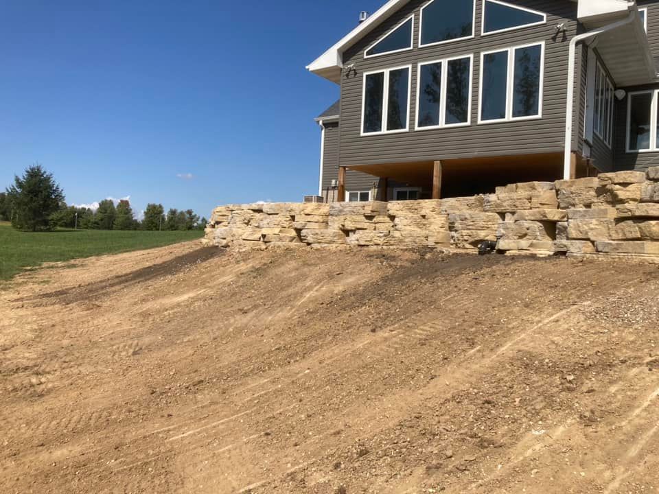 Stone retaining wall in front of a house on a hillside, with bare dirt and a blue sky.