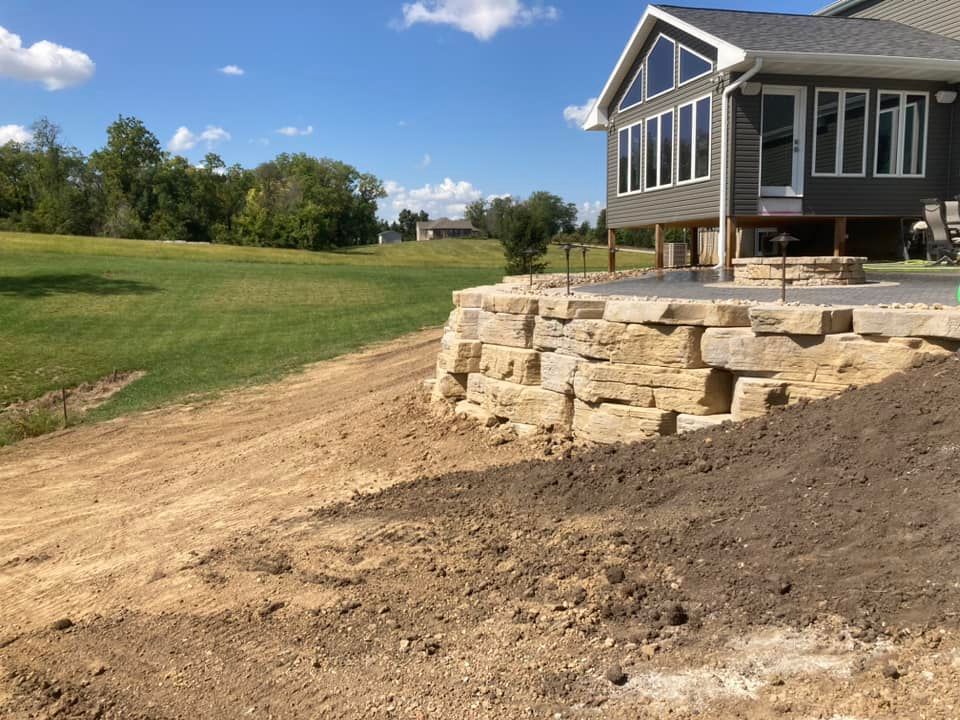 Stone retaining wall next to a grassy lawn and a house with large windows.