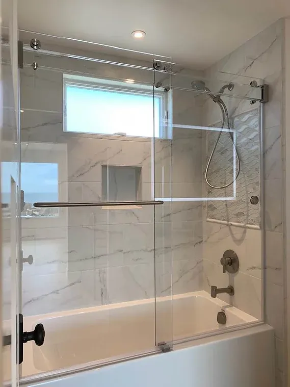 Bathtub with clear glass shower door, white tile wall, window, and silver shower fixtures.