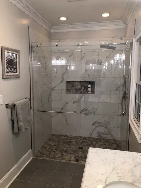Modern bathroom with a marble-tiled shower, glass door, and stone floor. A towel rack and small artwork are on the left wall.