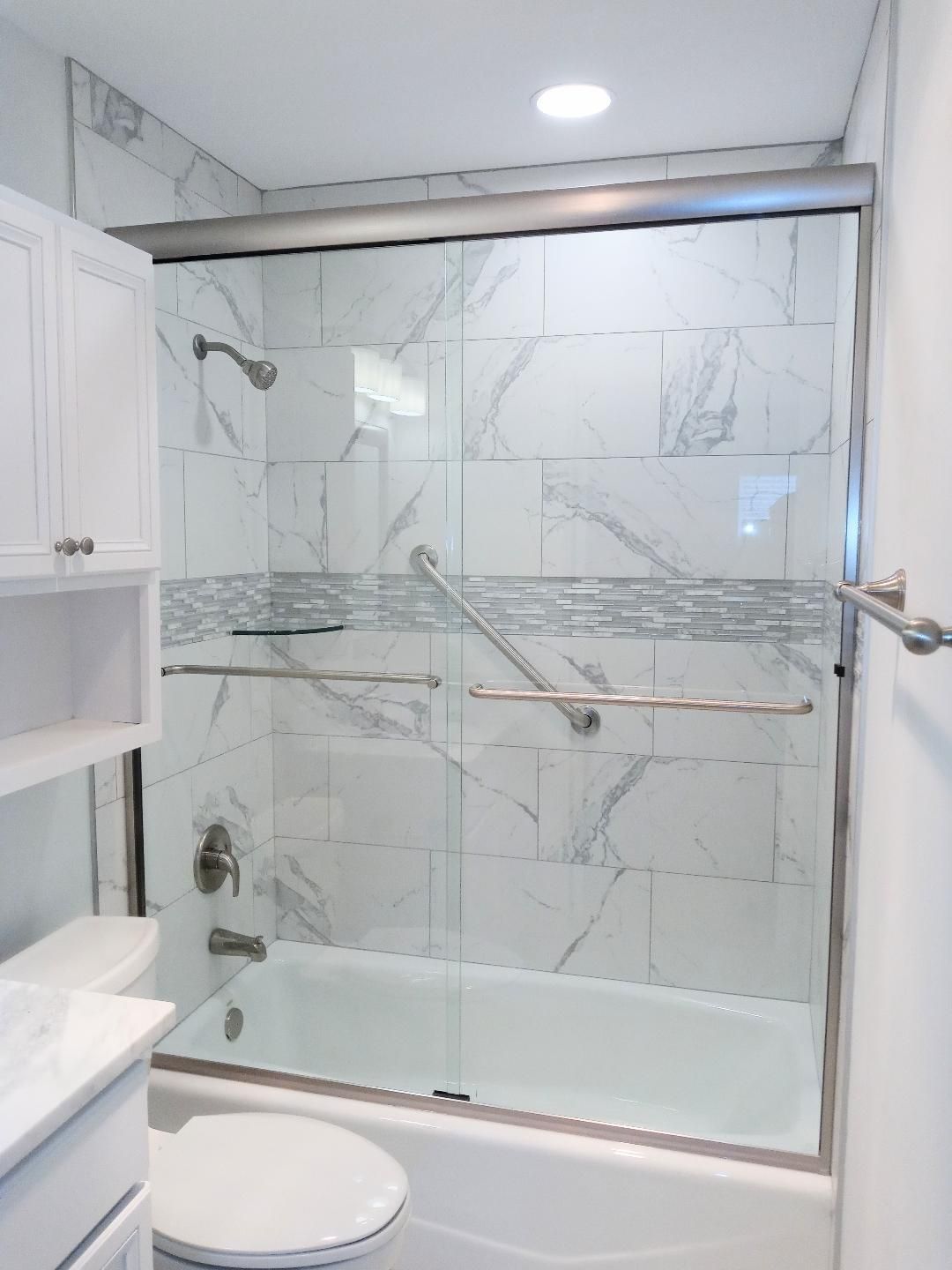 Bright bathroom with a white tub, marble-look tile, and a glass shower door. Includes a toilet, shelves.