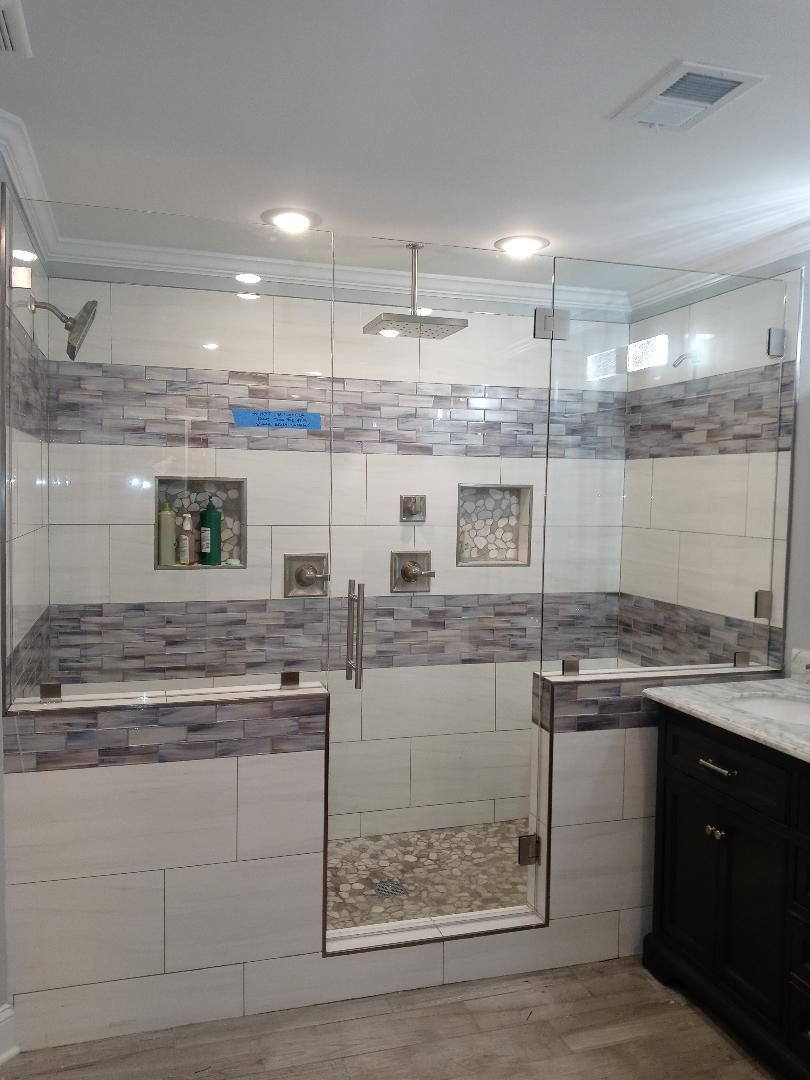 A newly renovated shower with glass doors and stone and white tile walls, including built-in shelving, a rain showerhead.