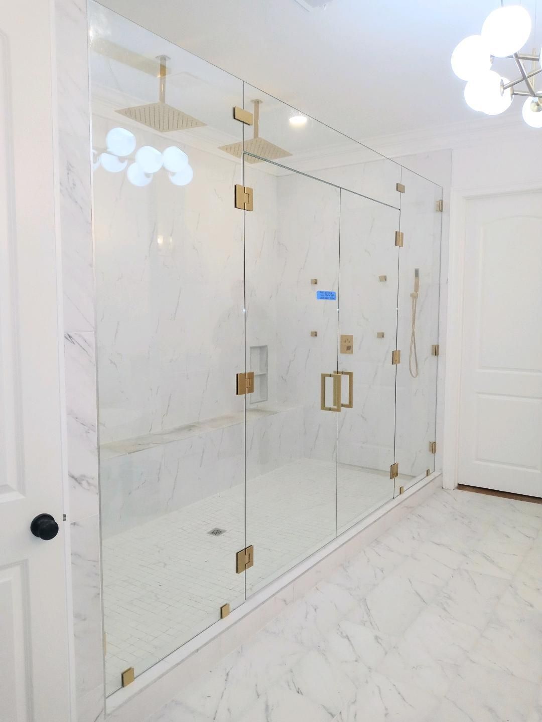 A luxurious shower with a glass enclosure, gold fixtures, and marble walls and floor. Bright white.