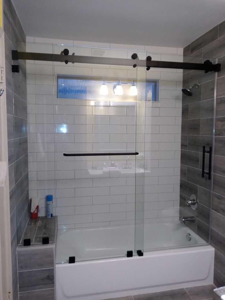 A modern bathroom with a sliding glass shower door over a white bathtub. Black hardware contrasts with the clear glass.
