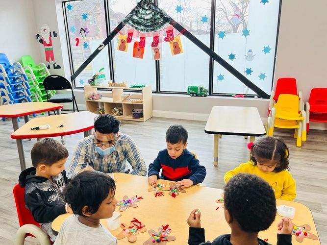 Children and teacher sit around a table, crafting. Inside a classroom with colorful chairs.