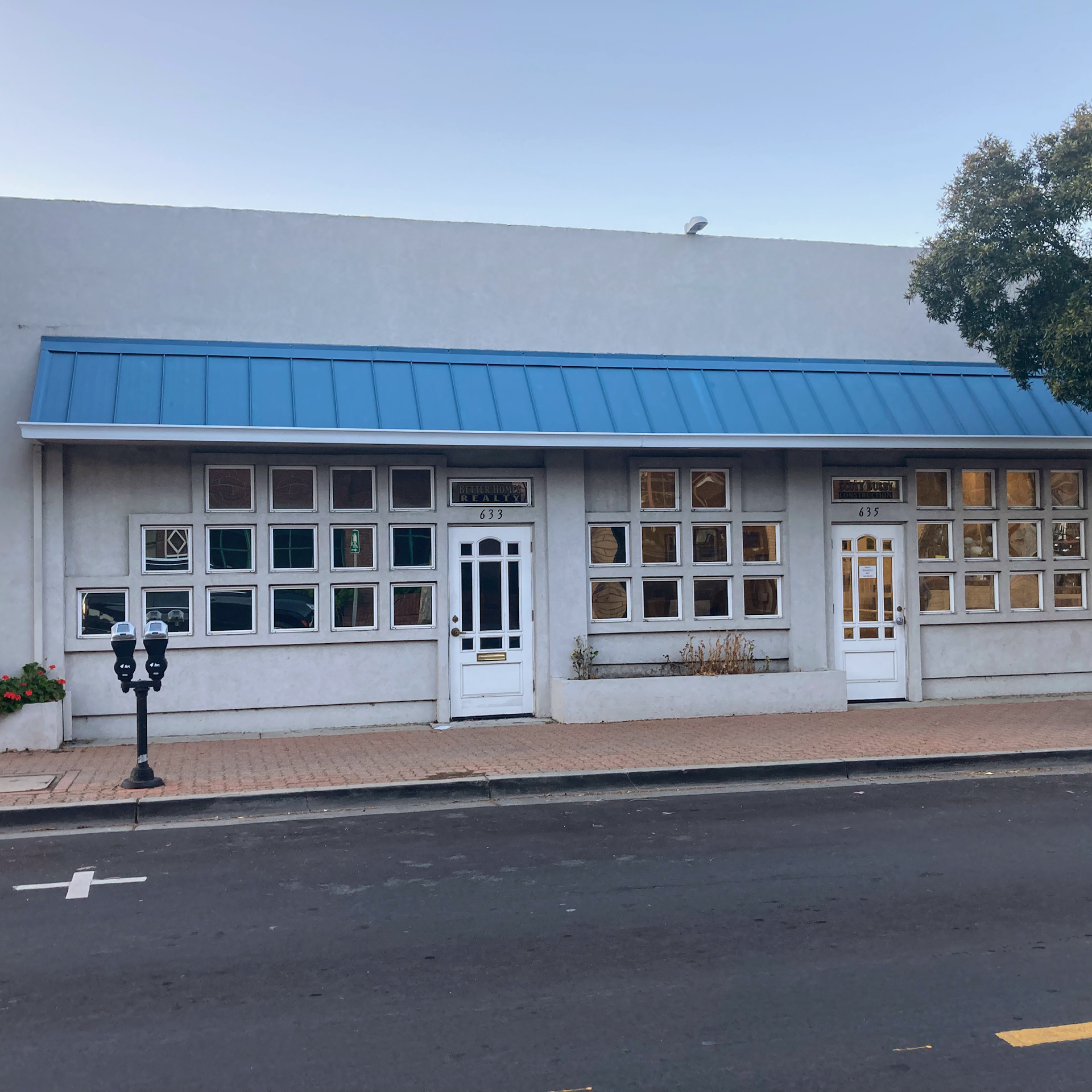 Single-story gray commercial building with a blue metal awning, grid-patterned windows, and two white entry doors.