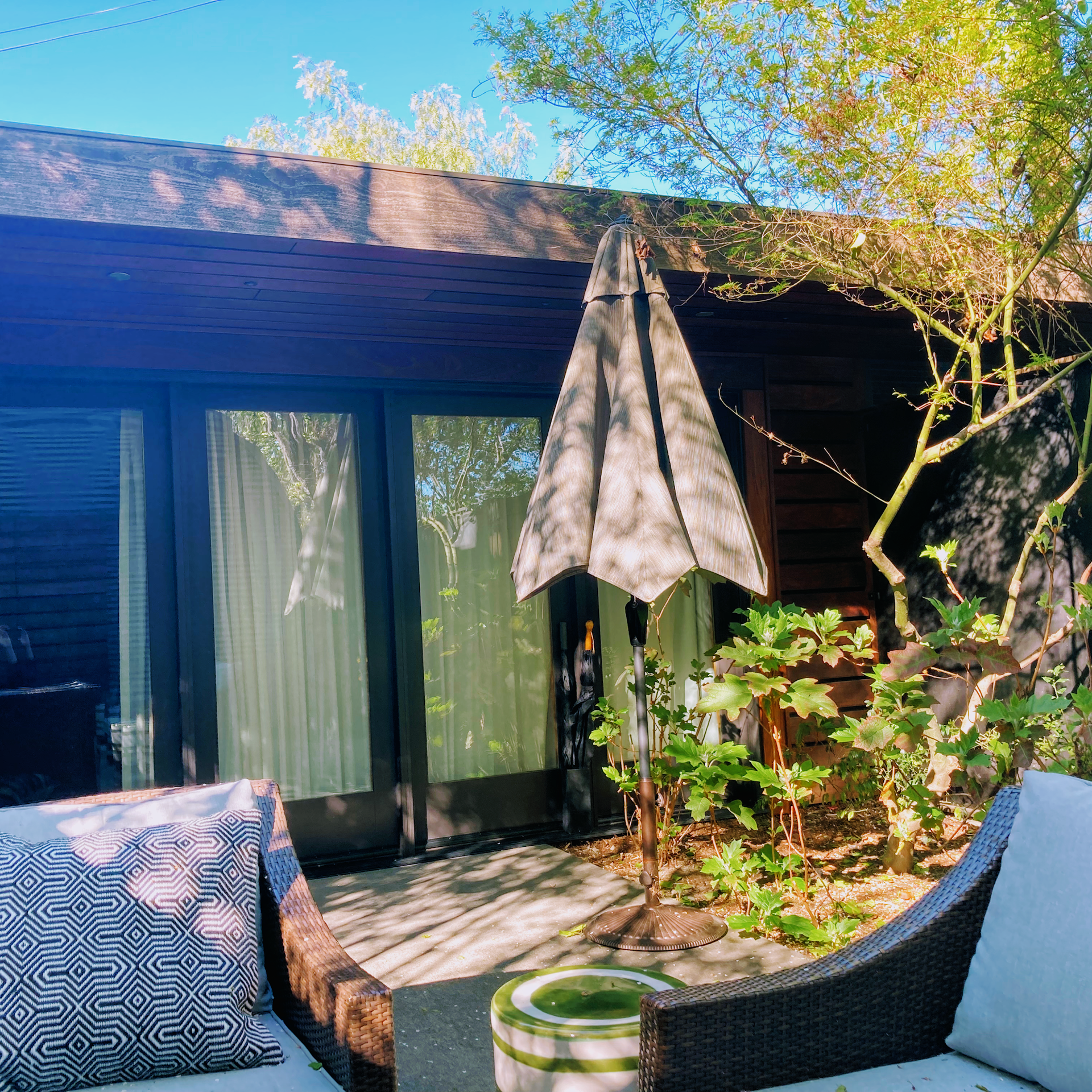 A patio area with two outdoor chairs, a small table, and a closed patio umbrella in front of a dark house with glass doors.