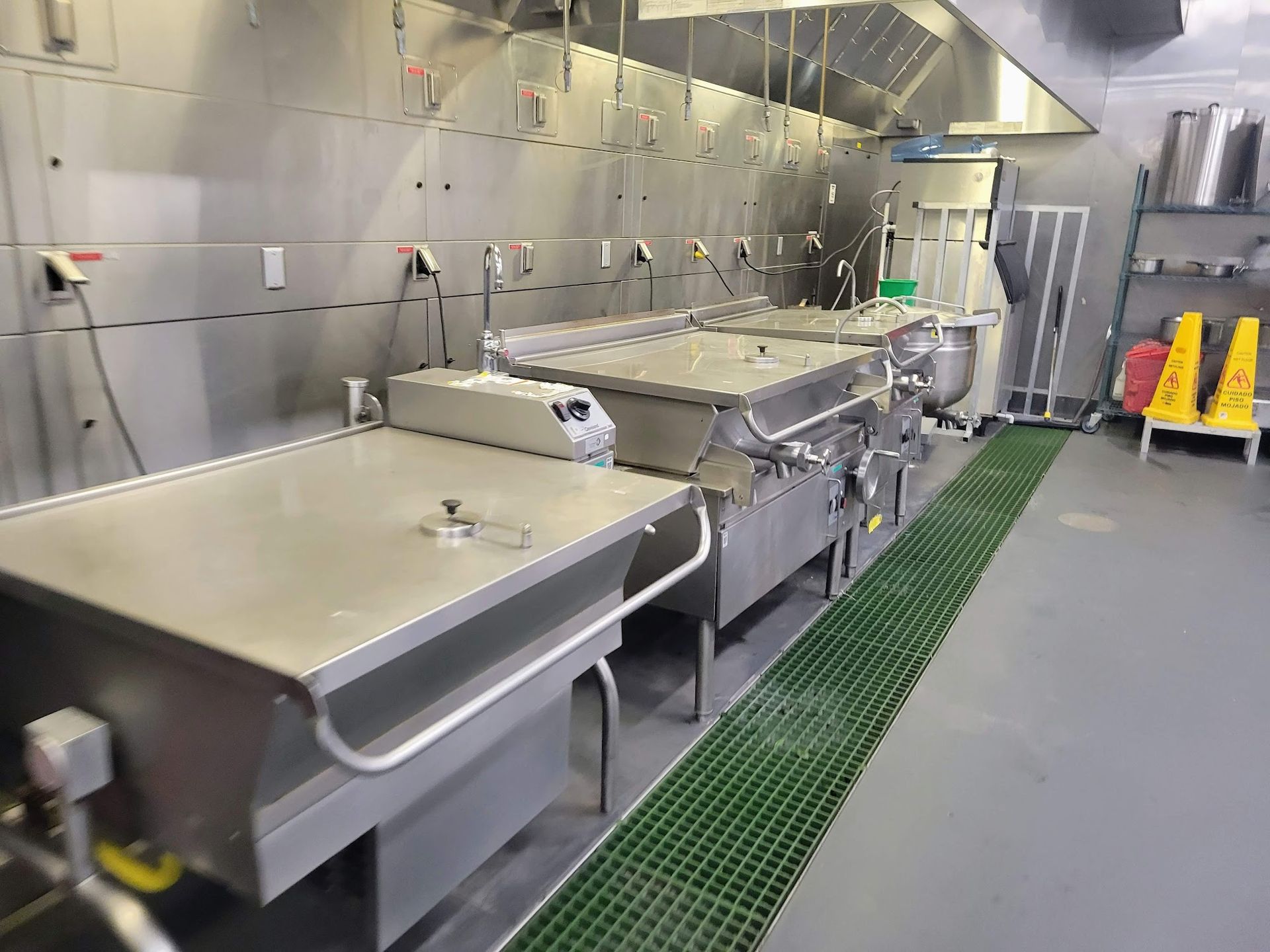 A row of industrial stainless steel tilting skillets in a professional commercial kitchen with a green floor mat.