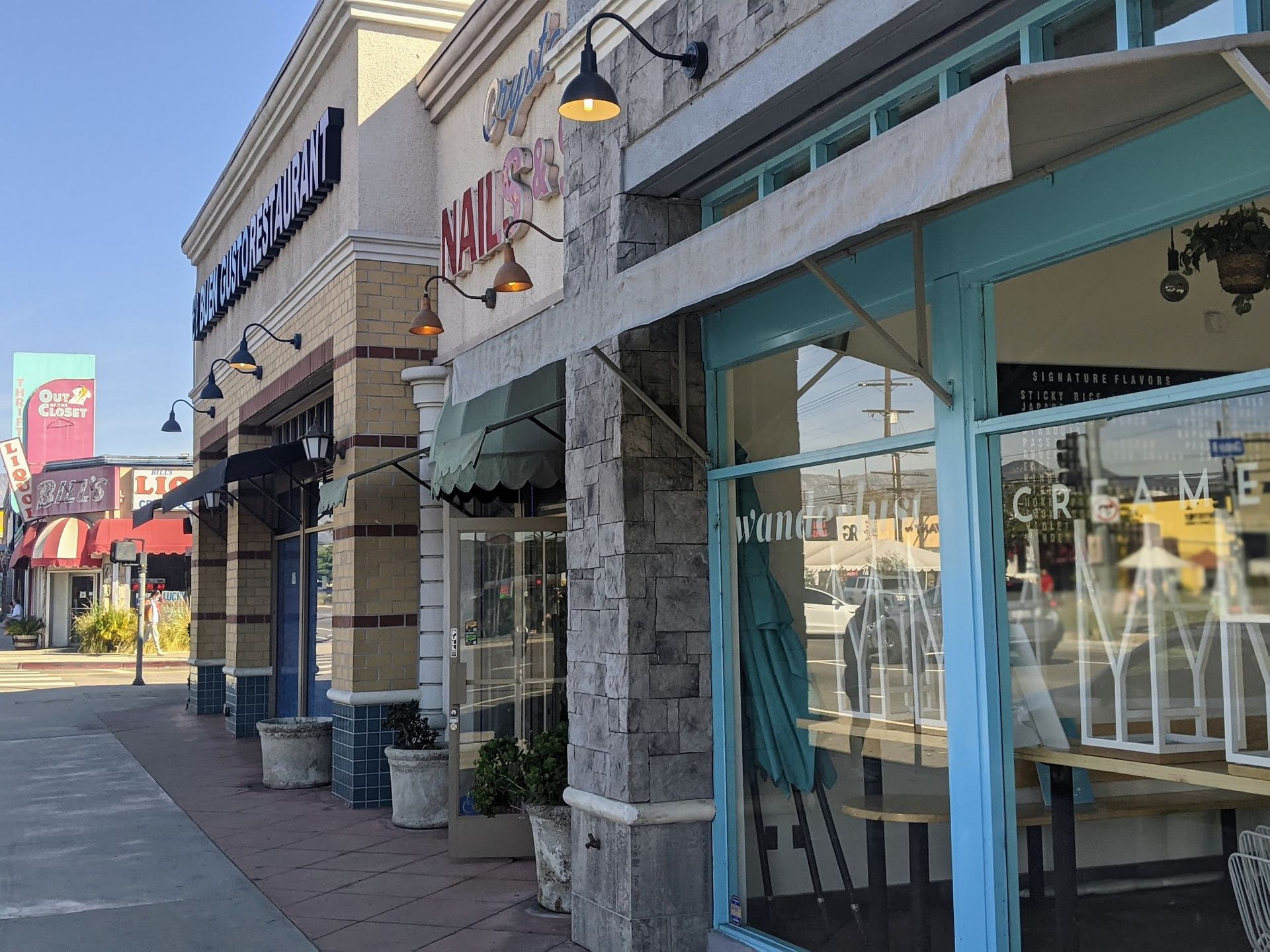 A sidewalk view of a commercial strip mall with light-colored brick facades and blue-framed storefront windows.
