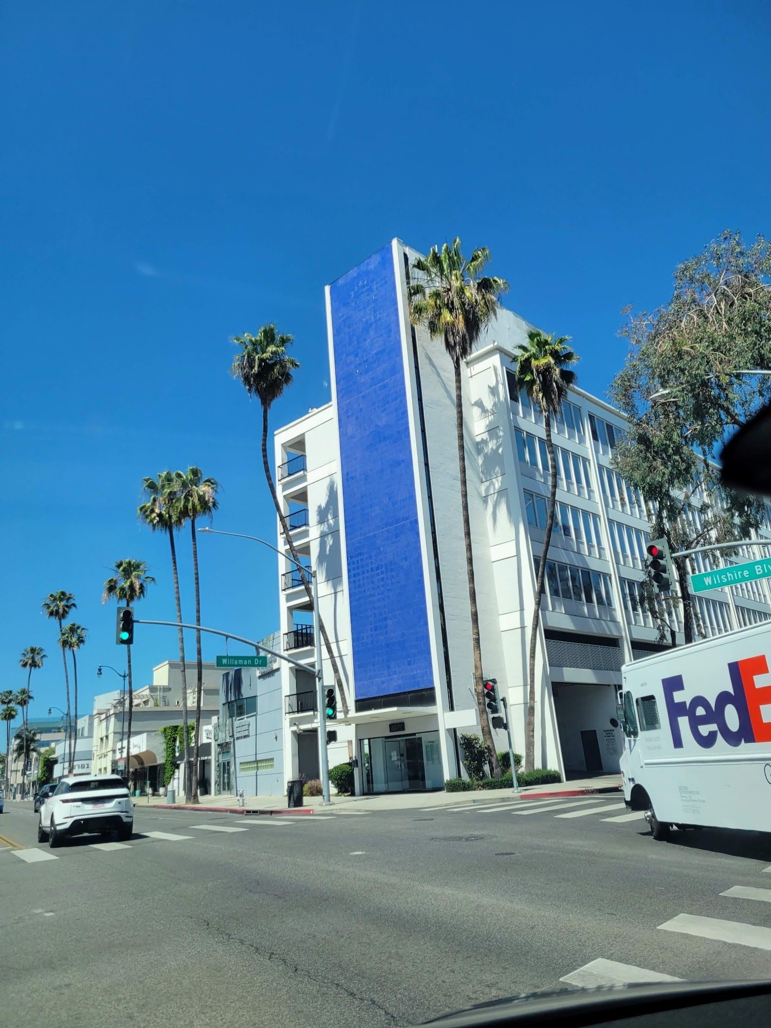 A white building with a vertical blue panel stands on a street corner lined with palm trees under a bright blue sky.