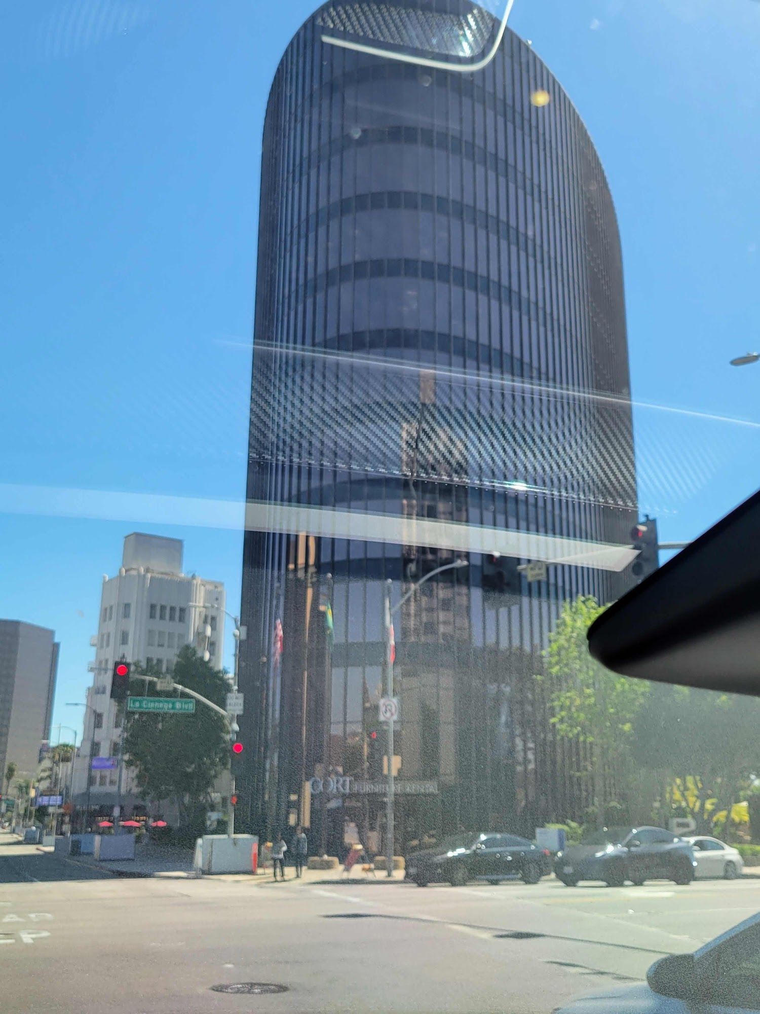 A tall, curved, dark glass skyscraper stands behind a street corner intersection under a clear blue sky.