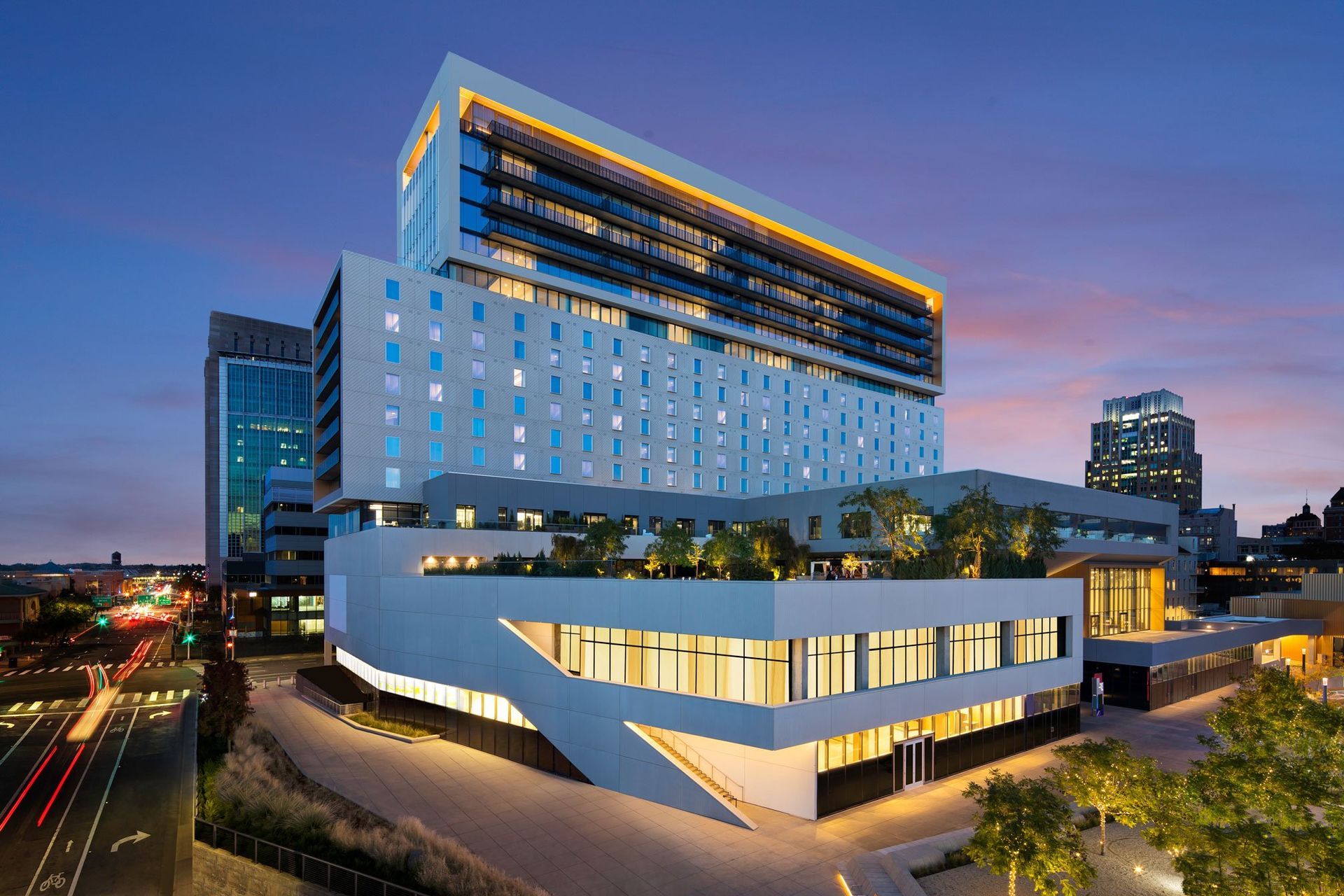A modern multi-story hospital building illuminated at dusk with a rooftop garden and light trails from traffic below.
