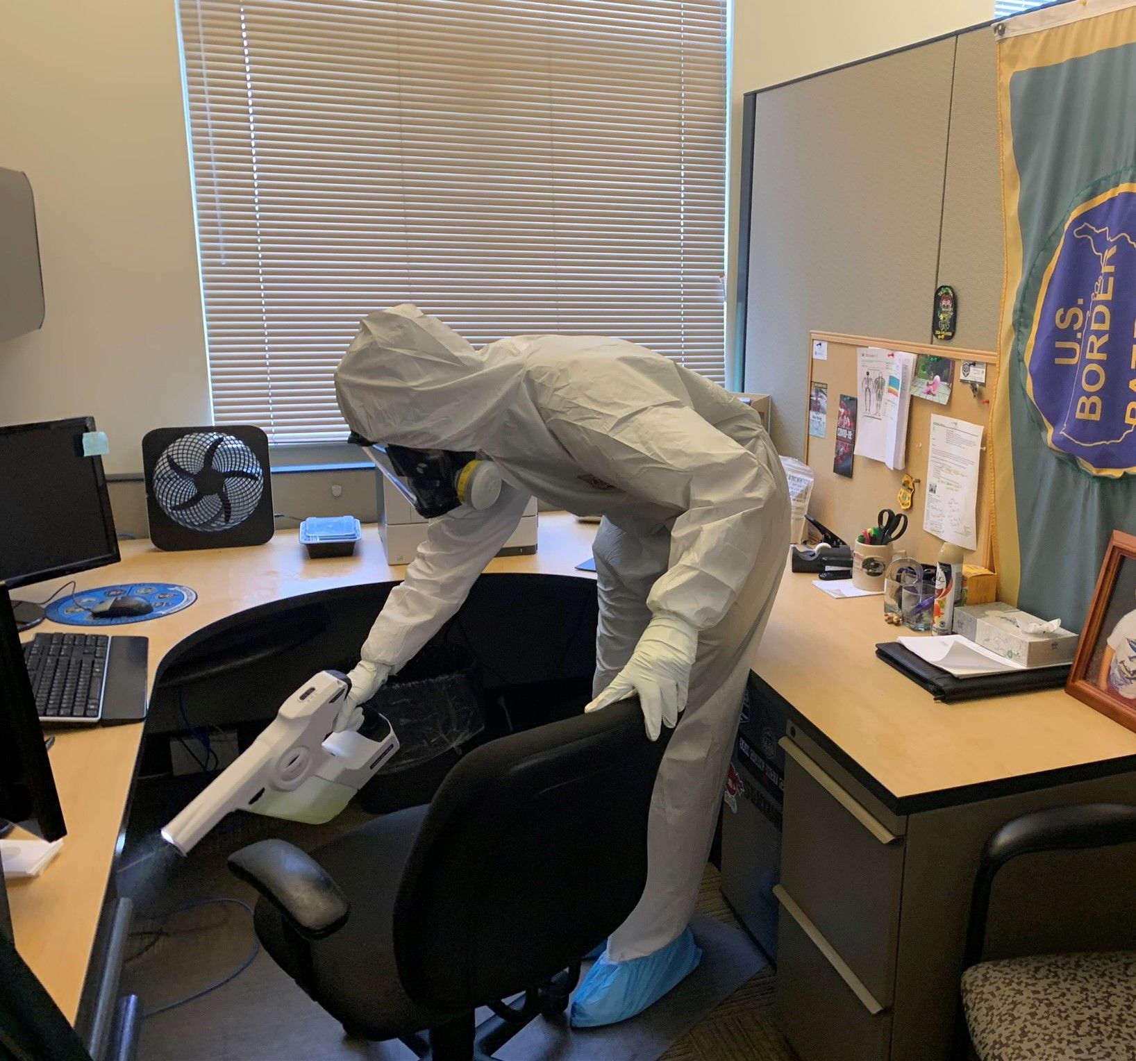 A person in a white hazmat suit and mask uses a handheld disinfectant sprayer to sanitize an office desk and chair.