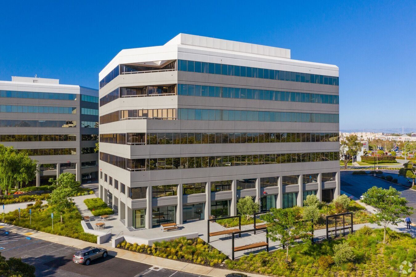 Modern mid-rise office building with glass windows, gray facade, and landscaped courtyard on a sunny day.