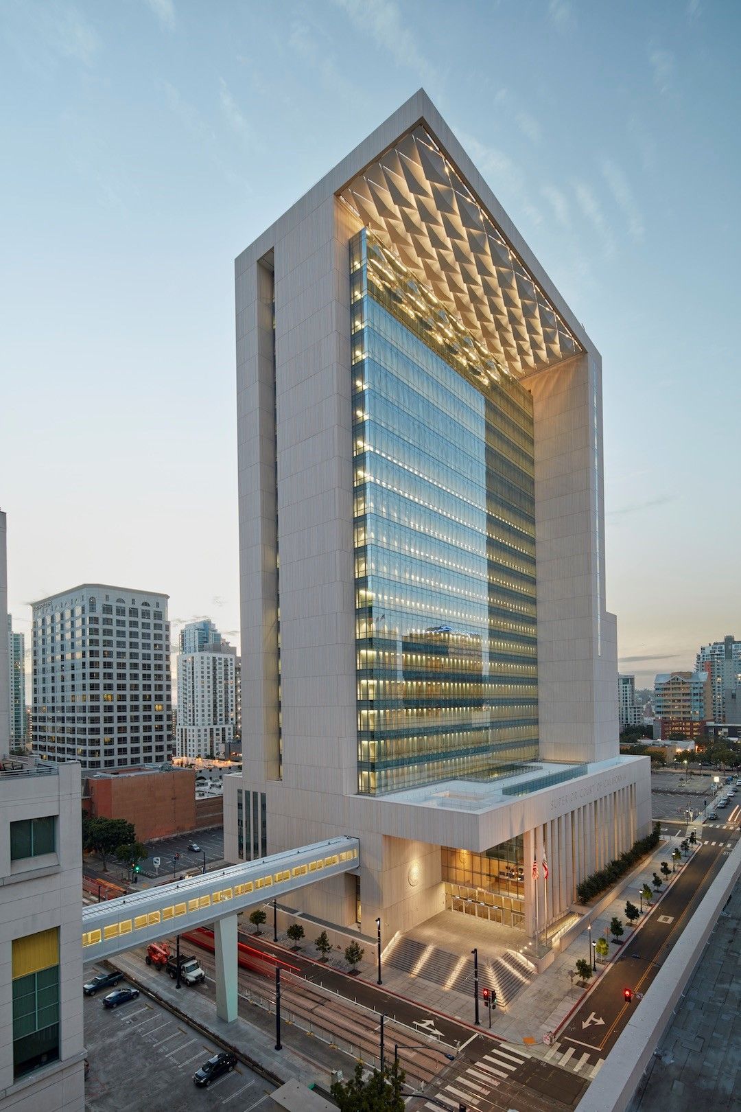 A tall, modern office building with a glass facade, a stone frame, and an elevated pedestrian bridge at sunset.