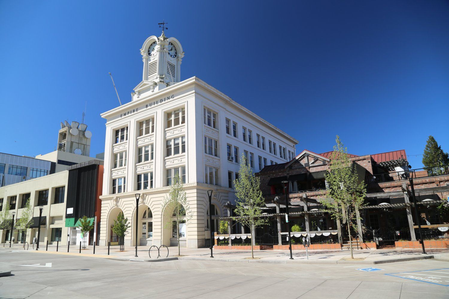 A white, four-story building with a clock tower stands on a city corner under a clear blue sky, next to a lower restaurant.
