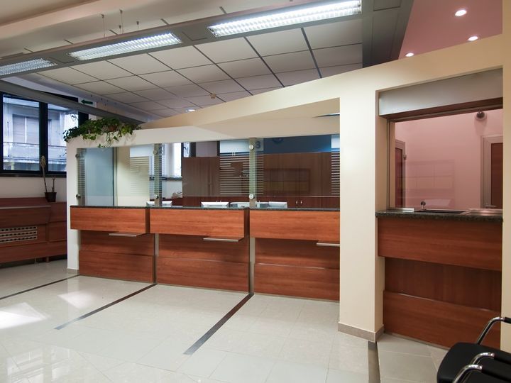 A modern office lobby featuring a long, wood-paneled reception desk, glass partitions, and white tile flooring.