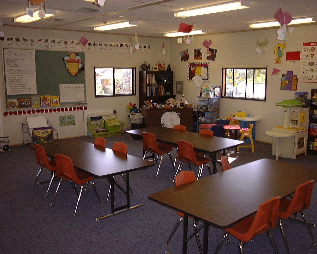 A classroom with dark tables and orange chairs, a chalkboard, windows, shelves, and various educational toys.