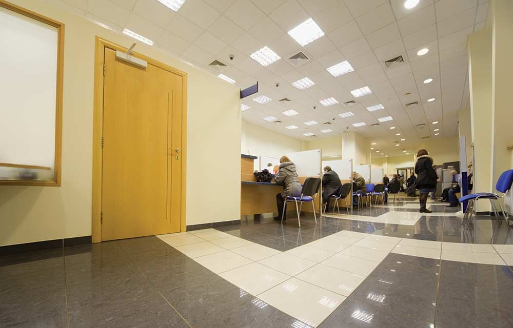 A bright, modern office lobby with light-colored walls, a wooden door, and people seated at individual service counters.