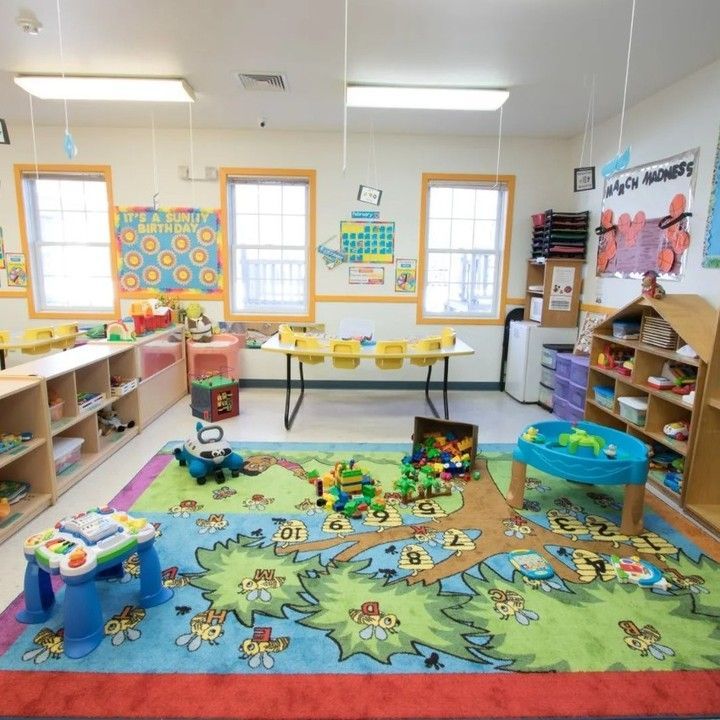 Brightly lit classroom with wooden shelves, child-sized tables, and a vibrant, tree-patterned area rug filled with toys.
