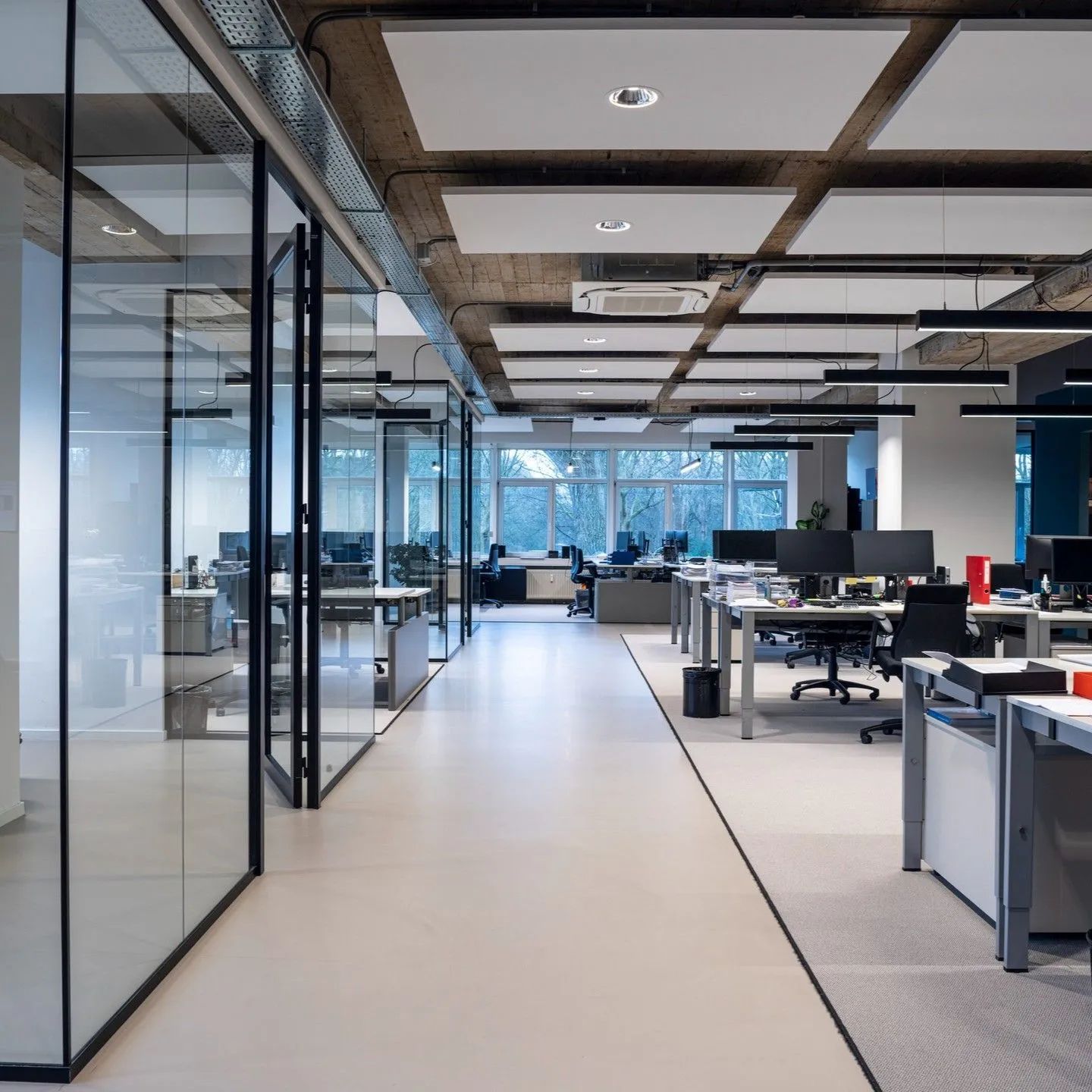 Modern open-plan office with glass walls, desks, monitors, and bright natural light