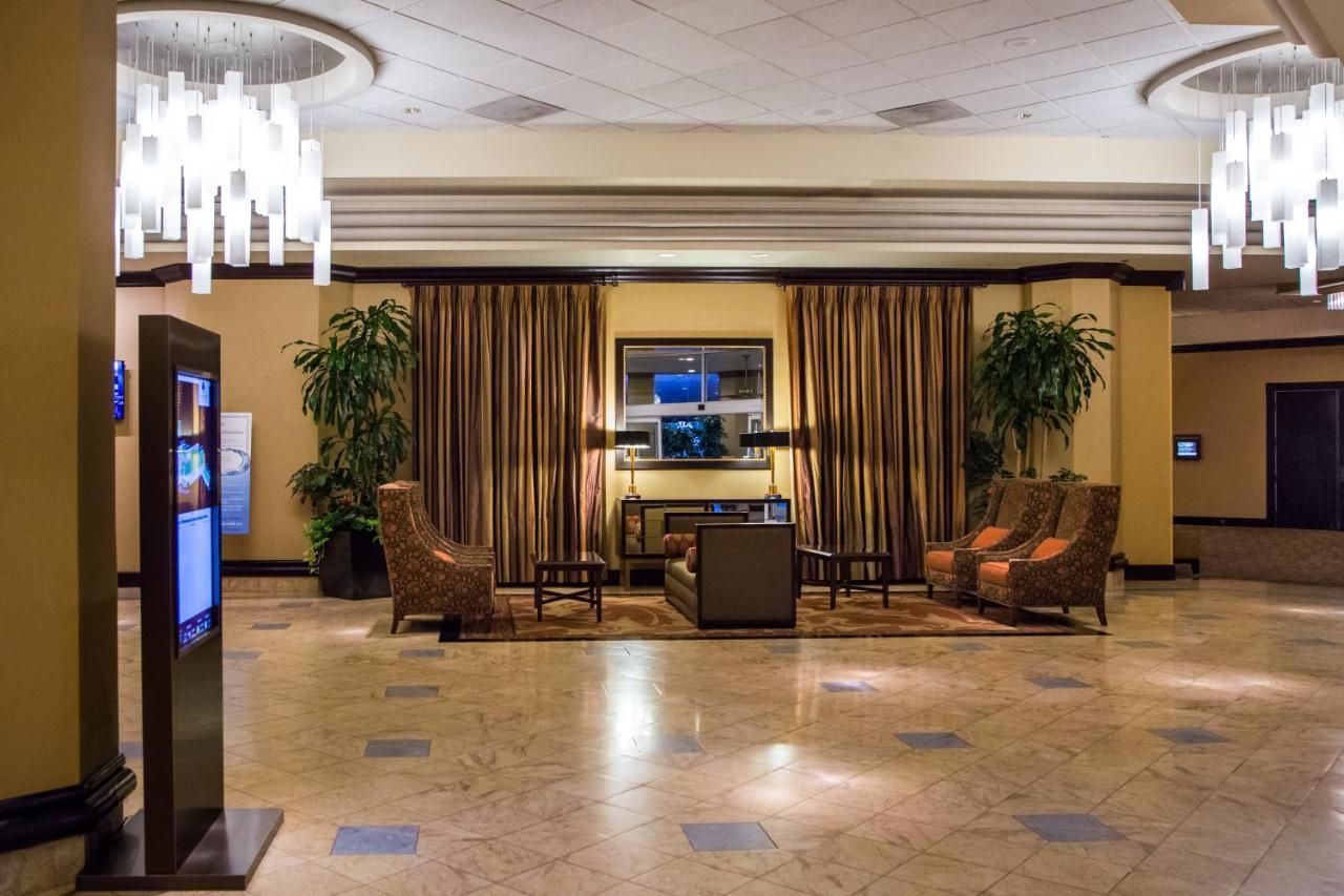 A hotel lobby with marble floors, patterned armchairs, potted plants, curtained walls, and two modern chandelier lights.