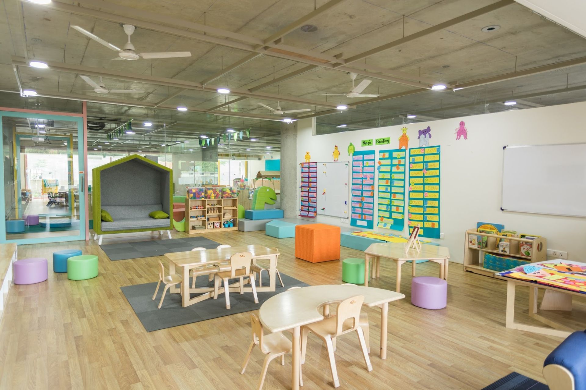 A bright, colorful preschool classroom with small tables, chairs, floor cushions, and a play nook on a wooden floor.