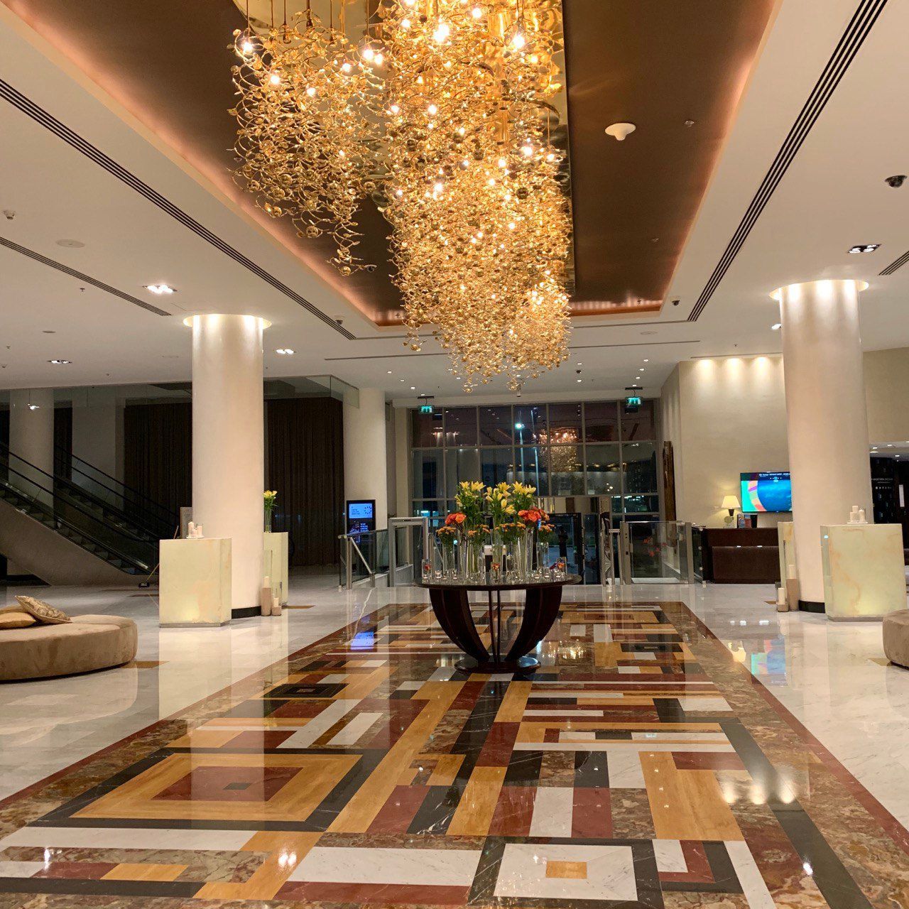 A luxury hotel lobby featuring a decorative crystal chandelier, a center table with flowers, and a geometric tiled floor.