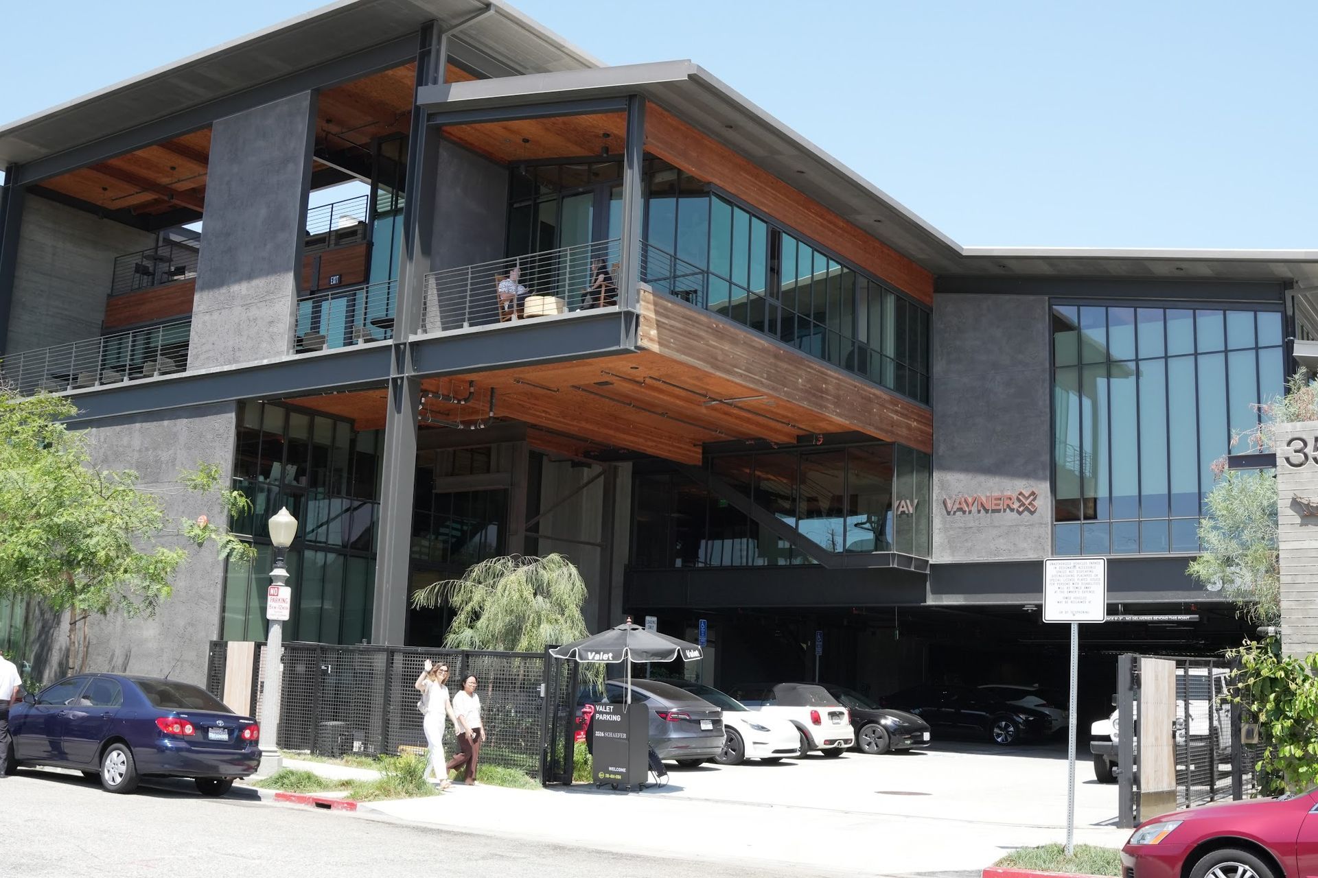 A modern two-story building with gray stone, steel beams, glass walls, and an open balcony above a parking level.