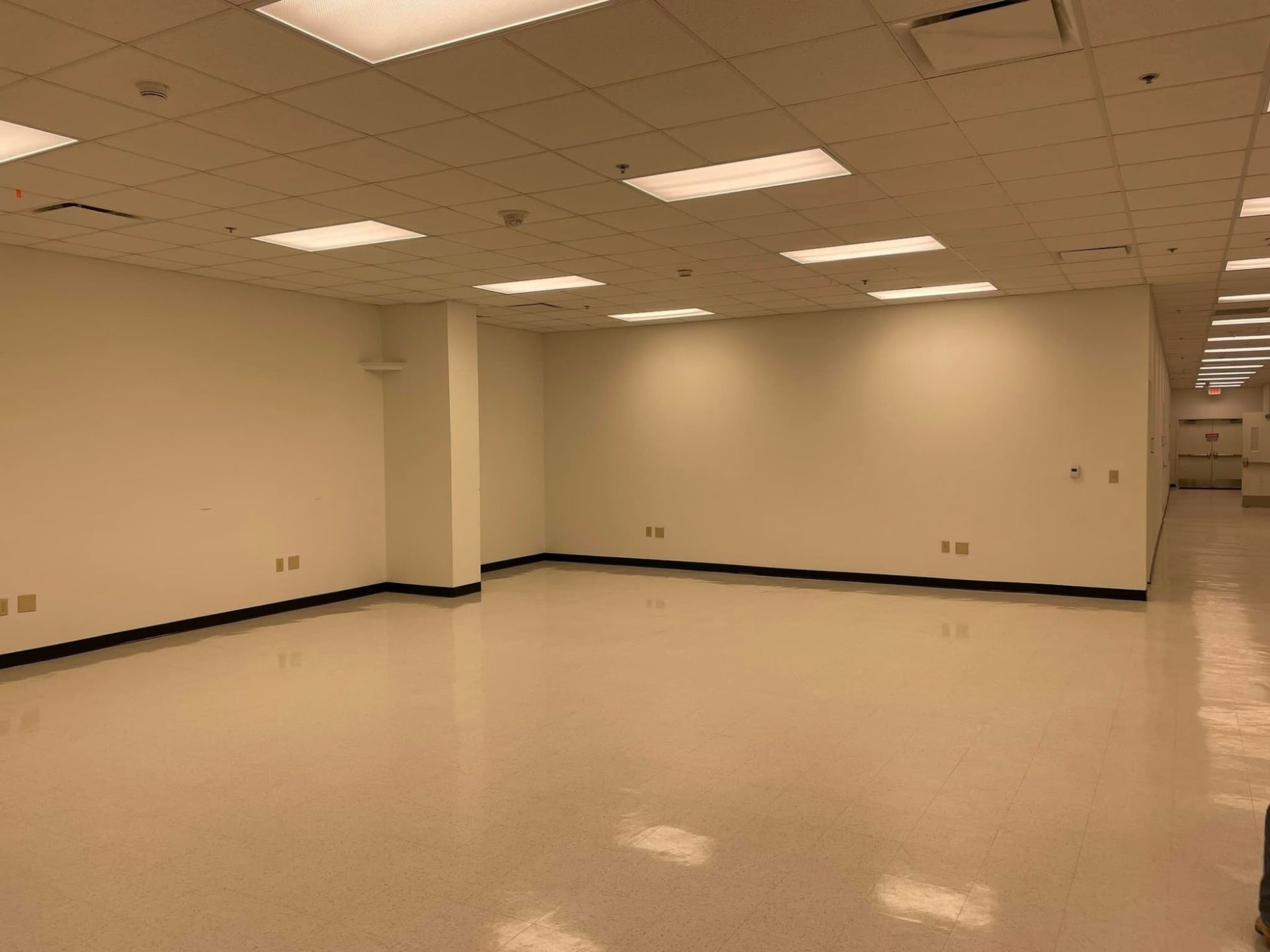 An empty, dimly lit office space with white walls, a drop ceiling with fluorescent lights, and glossy beige flooring.