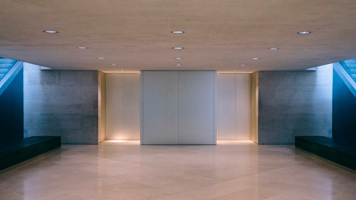 A minimalist, symmetrical indoor space featuring a smooth beige floor, two grey stone walls, and warm ambient lighting.