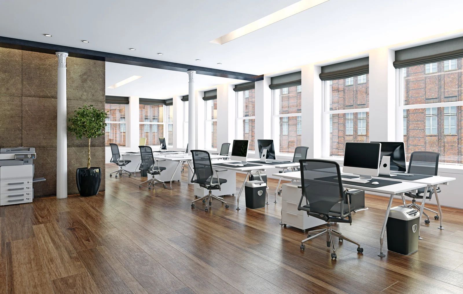 Modern open office with desks, computers, rolling chairs, and large windows overlooking brick buildings