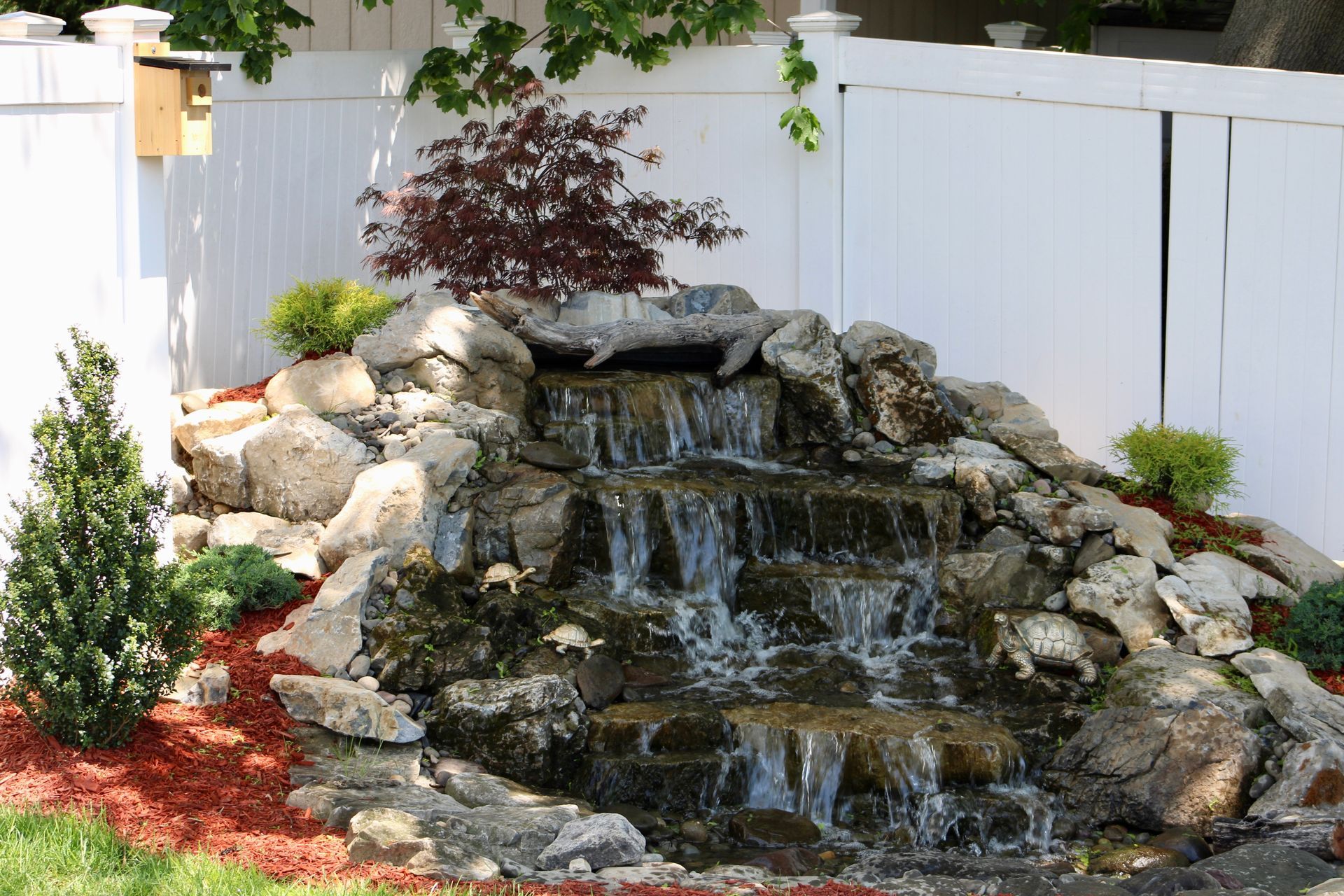 A waterfall in a backyard with a white fence in the background
